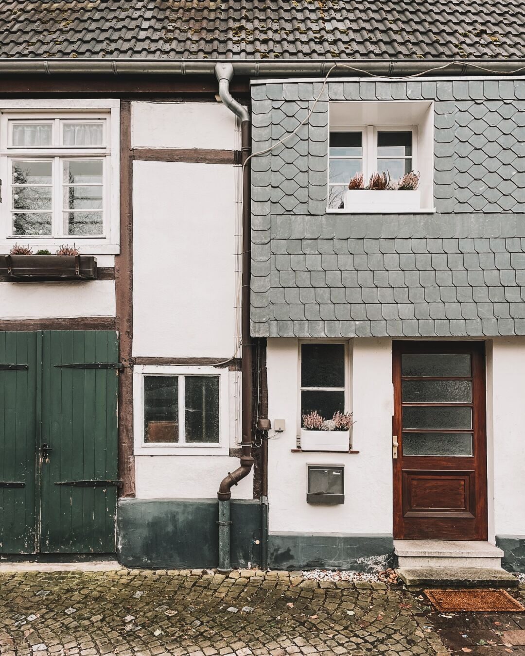 Die Fronten von zwei Fachwerkhäusern in Wiedenbrück. Man sieht jeweils nur die Hälfte. Weiße Fensterrahmen, eine grüne und eine braune Tür. In der Mitte sieht man ein Regenabwasserrohr. Auf den Fensterbänken des rechten Hauses stehen Blumen. Der Platz vor den Häusern ist aufwendig gepflastert.